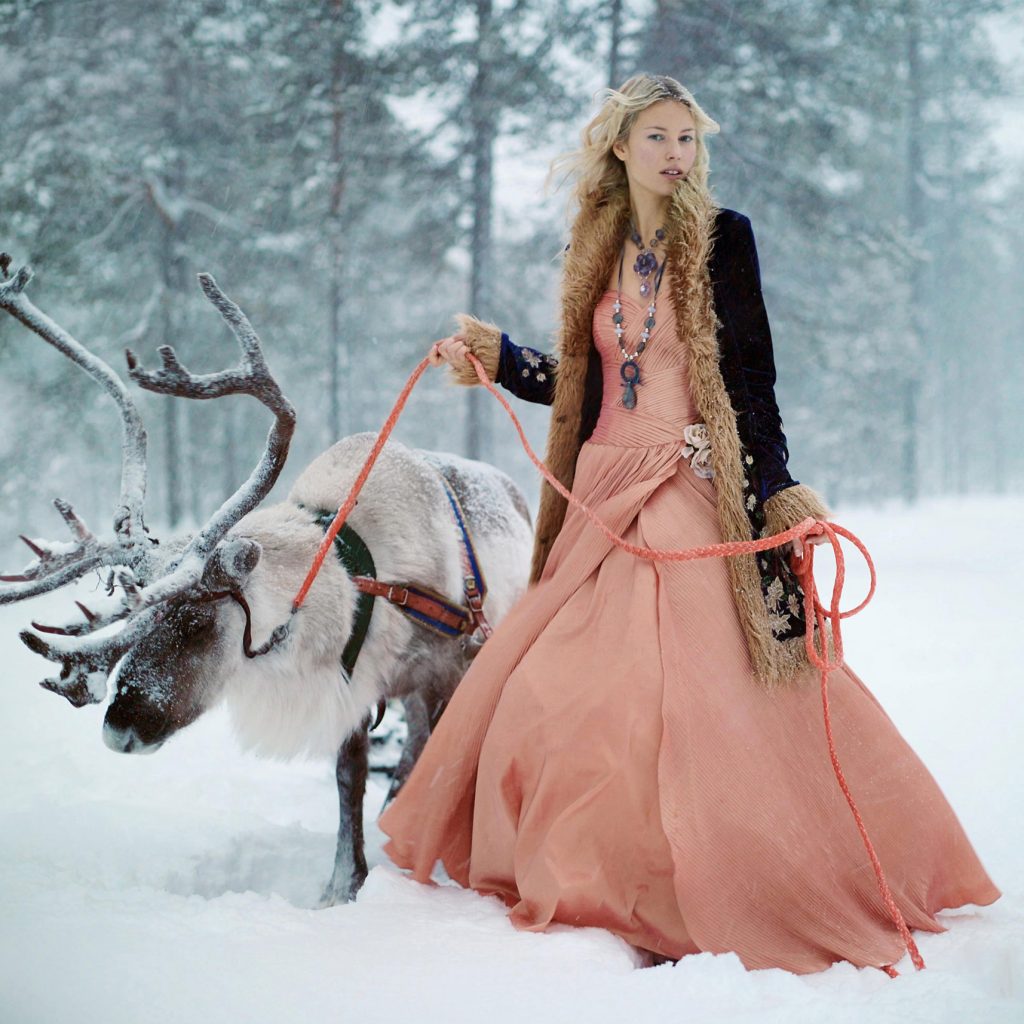 blond finnish model in lapland wearing peach long ball dress and winter sheepskin coat with reindeer on the leash standing in the snow with forest in the background